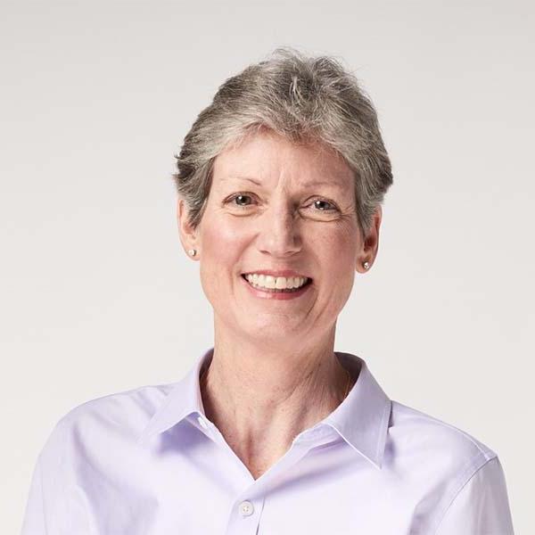 Professional portrait of a woman smiling, wearing a light purple blouse against a grey background.