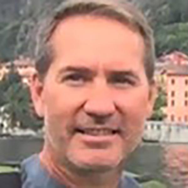 A smiling man with short hair posing for a picture with a scenic lake and mountains in the background. he's wearing a collared shirt and seems to be enjoying a lovely day outdoors.