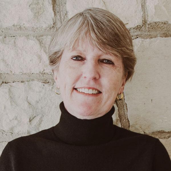 Portrait of a smiling woman wearing a black high-neck sweater and gold hoop earrings, standing against a brick background.